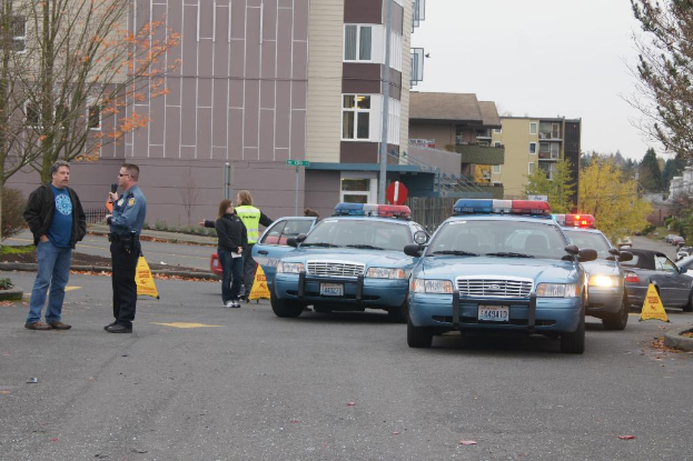 Autos auf einer Straße mit vier Personen in der Nähe, Gebäude mit Fenstern im Hintergrund, Bäume und Warnkeile.