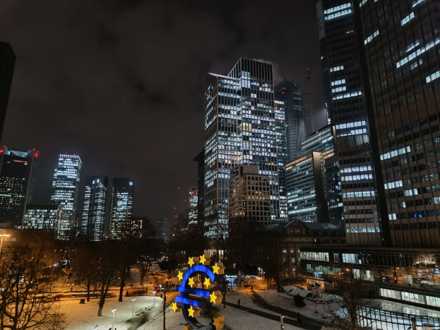 Europäischer Zentralbank-Hauptsitz in Frankfurt, Deutschland bei Nacht, mit beleuchteten Gebäuden, Bäumen, Straßenlaternen, Lichtern und Fußgängern unter bewölktem Himmel und einem Wasserzeichen.