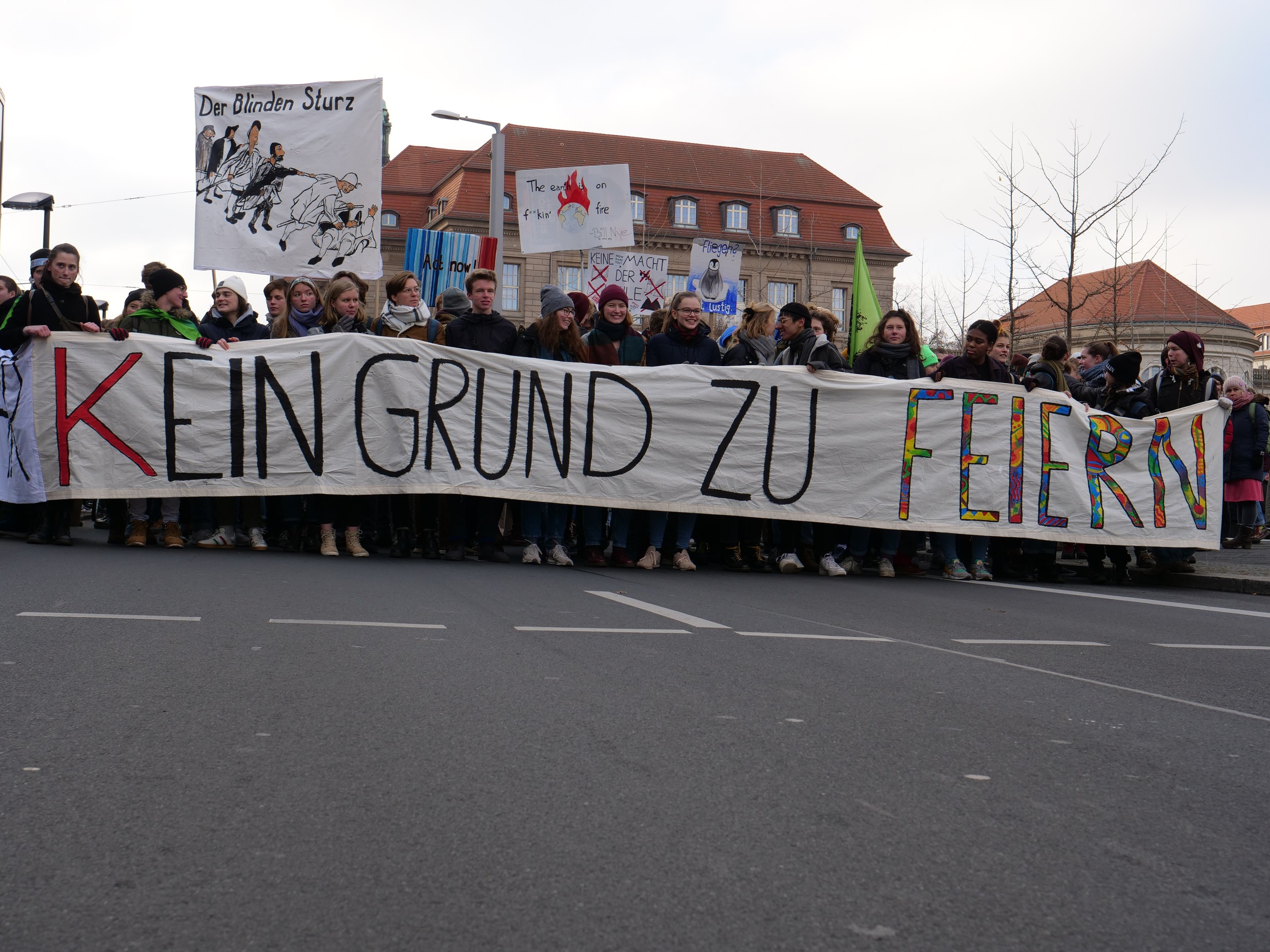 Demonstranten halten ein Transparent mit der Aufschrift "Kein Grund zu Feiern" auf einer Straße vor dem Hintergrund von Straßeninfrastruktur, Gebäuden, Bäumen und einem klaren Himmel.