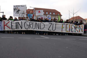 Demonstranten halten ein Transparent mit der Aufschrift "Kein Grund zu Feiern" auf einer Straße vor dem Hintergrund von Straßeninfrastruktur, Gebäuden, Bäumen und einem klaren Himmel.