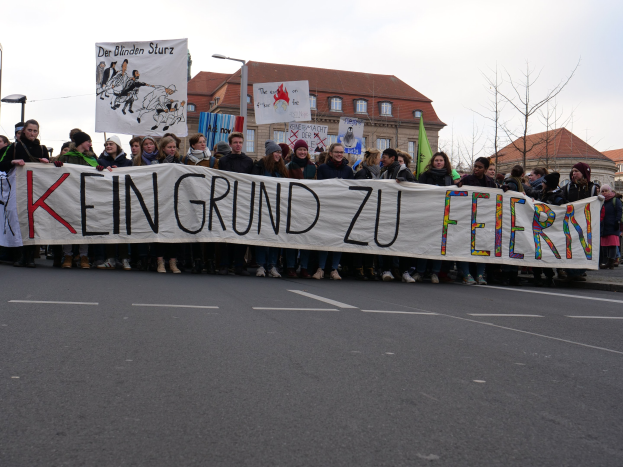 Demonstranten halten ein Transparent mit der Aufschrift "Kein Grund zu Feiern" auf einer Straße vor dem Hintergrund von Straßeninfrastruktur, Gebäuden, Bäumen und einem klaren Himmel.