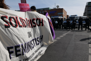 Gruppe von Menschen marschiert auf einer Straße und hält ein "Solidarität und Feminismus"-Schild, mit parkenden Fahrzeugen, Gebäuden, einer Schüsselantenne und einem klaren blauen Himmel im Hintergrund.
