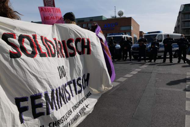 Gruppe von Menschen marschiert auf einer Straße und hält ein "Solidarität und Feminismus"-Schild, mit parkenden Fahrzeugen, Gebäuden, einer Schüsselantenne und einem klaren blauen Himmel im Hintergrund.
