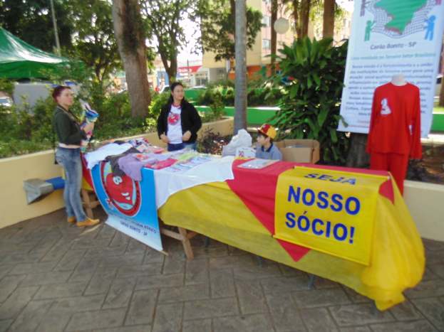 Zwei Frauen und ein Junge vor einem Tisch mit einer Decke und einem Banner, auf dem Kleidung liegt, mit Bäumen, einem Gebäude und einer Schaufensterpuppe mit einem Banner im Hintergrund.