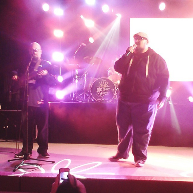 Zwei Männer auf der Bühne, einer hält eine Gitarre und der andere ein Mikrofon, mit einer Person, die ein Handy in der Hand hält, im Vordergrund und einem Drumset und Scheinwerfern im Hintergrund.