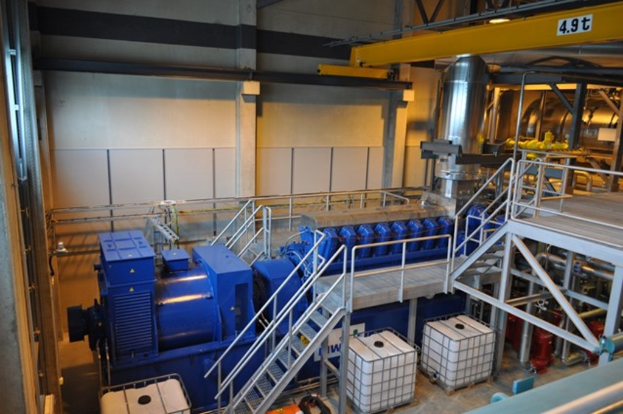 Ein Fabrikinnere mit Trägern, einer Treppe, Geländer, Containern, Lichtern, Rohren und Wänden.