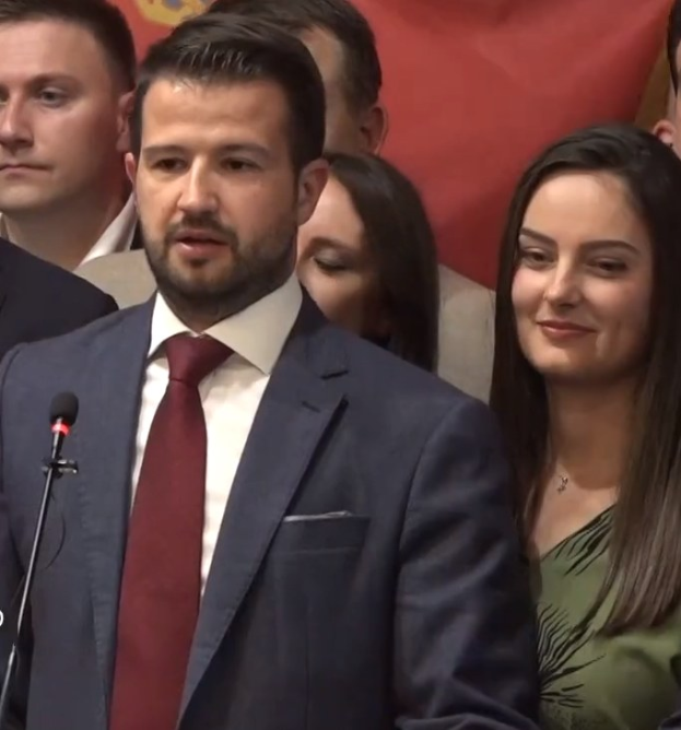 Ein Mann im Anzug spricht in ein Mikrofon an einem Rednerpult, mit einem Banner im Hintergrund und einer Menge davor.