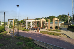 Ein Parkweg mit Holzpfählen, Bänken, Pflanzen, Gras, Steinen, Straßenpfählen, Straßenlaternen, Strommasten mit Drähten, Bäumen, Gebäuden und einem Turm unter einem klaren blauen Himmel.