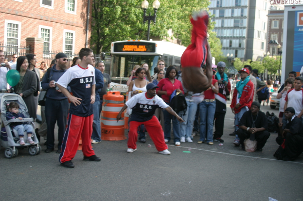 Eine volle Szene mit Menschen, einige tragen Mützen, ein Kind in einem Kinderwagen, ein Ballon und ein Verkehrshindernis; ein Bus, Bäume, Gebäude mit Fenstern, ein Laternenmast und ein Geländer sind im Hintergrund zu sehen.