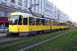 Eine gelbe und weiße Straßenbahn mit Menschen darin f√§hrt auf Schienen neben hohen Geb√§uden, mit Gras auf der rechten Seite, Autos auf der linken Seite und einer Kulisse aus Geb√§uden, B√§umen, Masten, Dr√∂hen und Himmel.