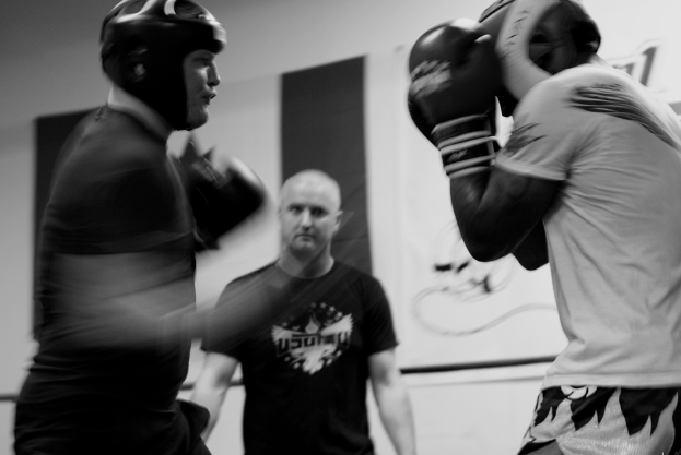 Zwei Boxer in Helm und Handschuhen k├Ąmpfen im Ring, mit einem Schiedsrichter dazwischen und einem Banner an der Wand im Hintergrund.