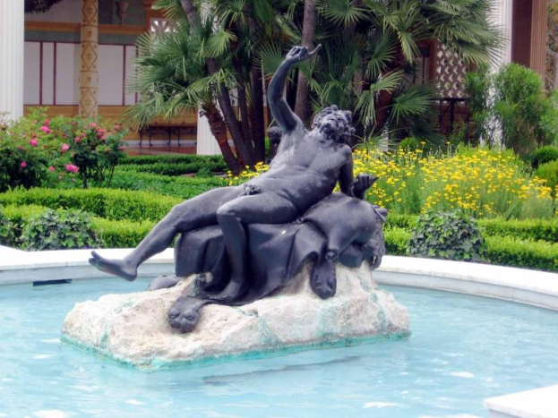 Ein Pool mit einer Skulptur in der Mitte, umgeben von Pflanzen, Bäumen und Säulen im Hintergrund.