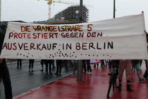 Eine Gruppe von Menschen steht auf einer Stra√e und hölt ein Banner mit der Aufschrift "Die Wrangellstra√e 5 Protestiert Gegen den Ausverkauf in Berlin", mit Fahrrödern, Masten, Gebäuden und einem Kran im Hintergrund und einem sichtbaren Himmel.