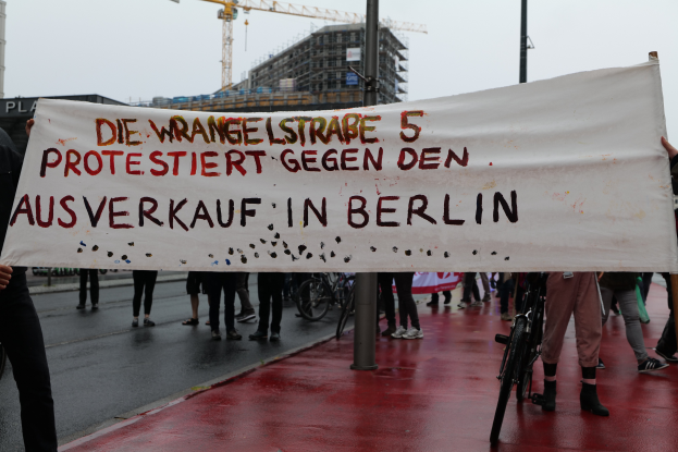 Eine Gruppe von Menschen steht auf einer Stra√e und hölt ein Banner mit der Aufschrift "Die Wrangellstra√e 5 Protestiert Gegen den Ausverkauf in Berlin", mit Fahrrödern, Masten, Gebäuden und einem Kran im Hintergrund und einem sichtbaren Himmel.