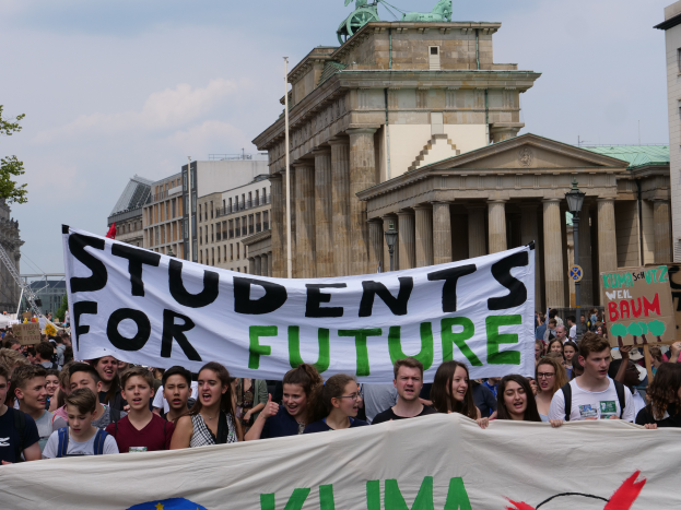 Eine Gruppe von Studenten marschiert in Berlin, mit einem bunt bemalten Banner, auf dem "Students for Future" steht, im Hintergrund Gebäude, Bäume und Himmel.