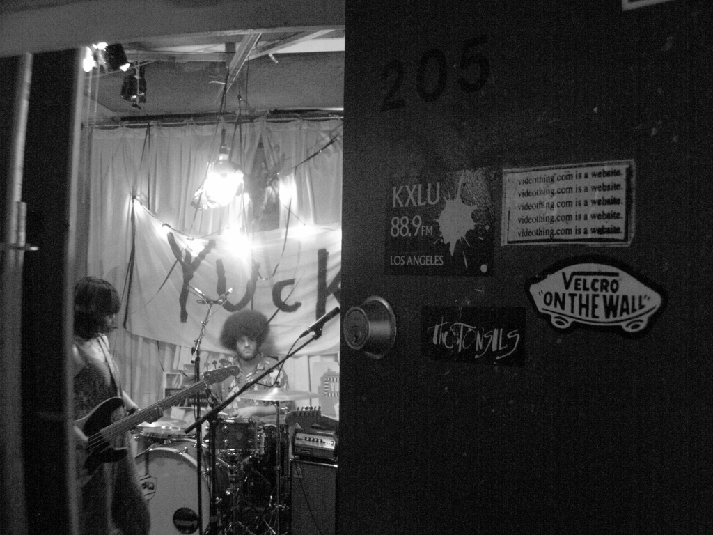 Ein Schwarz-Weiß-Bild eines Mannes, der vor Trommeln sitzt, einer Frau, die Gitarre spielt, und einem Banner mit der Aufschrift 'YUCK' im Hintergrund, mit einer sichtbaren Tür.