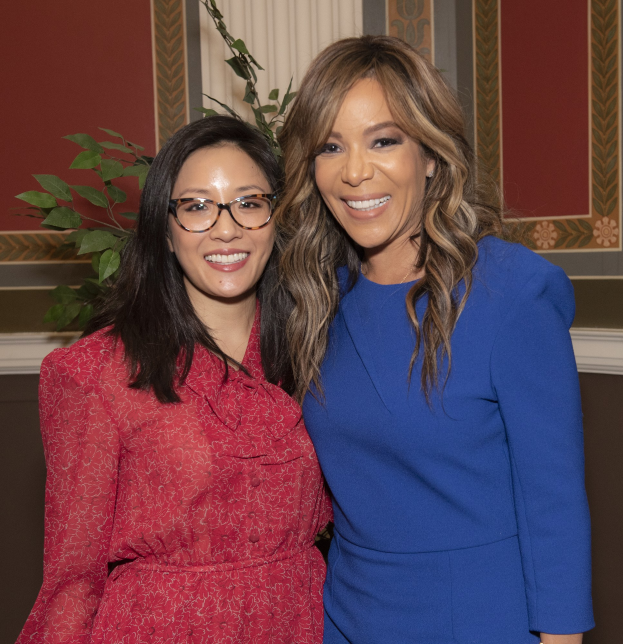 Zwei Frauen, die auf der 2019 Women in Film Pre-Oscar-Luncheon zusammen lächeln, eine in einem blauen Kleid und die andere in einem roten Kleid mit Brille, stehen neben einer Pflanze und einem Vorhang.