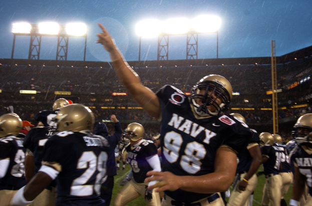Navy-Football-Spieler in Marineblau und Gold feiern ein Touchdown auf einem regennassen Rasenfeld, mit Stadionbeleuchtung und Zuschauern im Hintergrund.