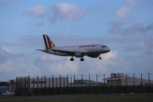 Germanwings Airbus A320-200 beim Start am Frankfurter Flughafen mit Gras darunter, einem Zaun im Vordergrund, Gebäuden und Bäumen im Hintergrund und einer bewölkten Himmel