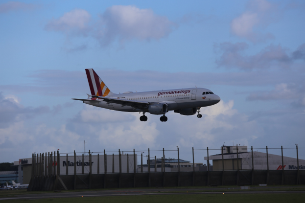 Germanwings Airbus A320-200 beim Start am Frankfurter Flughafen mit Gras darunter, einem Zaun im Vordergrund, Gebäuden und Bäumen im Hintergrund und einer bewölkten Himmel