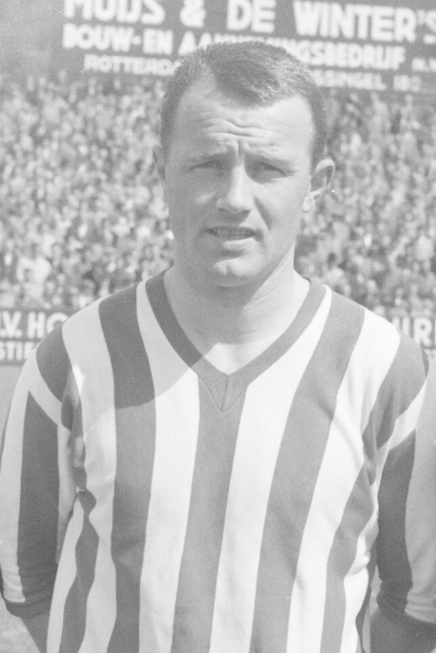 Ein Schwarz-Weiß-Foto eines Mannes in einem gestreiften Hemd, der vor einer Menge steht, mit einer Tafel im Hintergrund, der legendäre niederländische Fußballspieler Bertrand.
