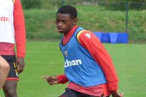 Ein junger Mann in einem blauen T-Shirt und schwarzen Shorts spielt Fußball auf einem grünen Rasen, der von einem Zaun, Pflanzen und Bäumen umgeben ist, während eines Trainings.