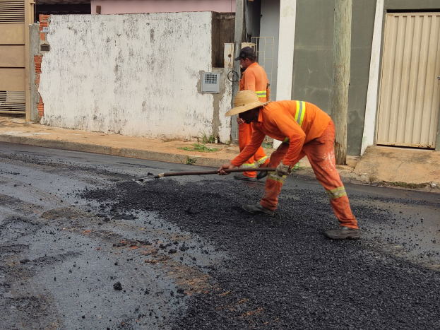 Zwei Männer in orangefarbenen Overalls und Hüten arbeiten an einer Straße, wobei einer ein Objekt hält, vor Gebäuden mit Läden.