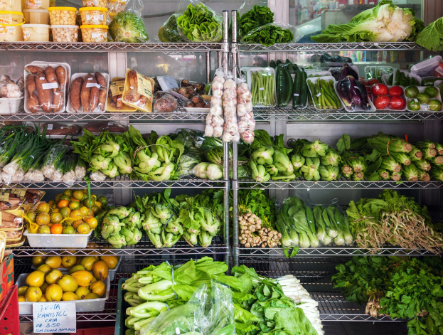 Ein Gang in einem Supermarkt mit frischem Gemüse und Obst, verpackten Artikeln, Kartons, einer Schautafel und einem Glasfenster im Hintergrund.