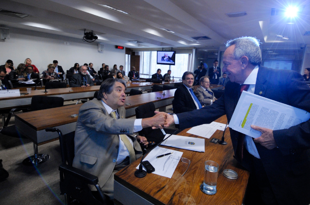 Eine Gruppe von Menschen ist in ein Gespräch mit ein paar im Hintergrund sitzenden Individuen vertieft, unter einem Dach mit Beleuchtungskörpern, wie auf einem Fernsehbildschirm zu sehen.