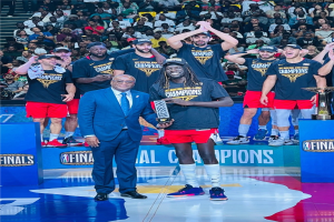 Eine Gruppe von Männern auf einem Basketballfeld, die Trophäen halten, mit Zuschauern im Hintergrund und zwei Trophäen auf Podesten rechts.