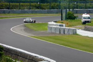 Ein Rennwagen fährt auf einer Rennstrecke mit einem Polizeiwagen im Hintergrund, umgeben von Grünfläche und Absperrungen, mit Text am unteren Bildrand.