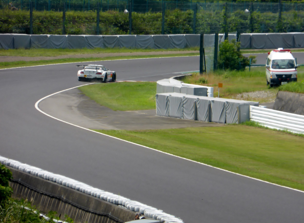 Ein Rennwagen fährt auf einer Rennstrecke, im Hintergrund ist ein Polizeiwagen zu sehen, umgeben von Gras, Pflanzen, Bäumen und einer Umzäunung, mit einem Fahrzeug auf der rechten Seite und Text unten.