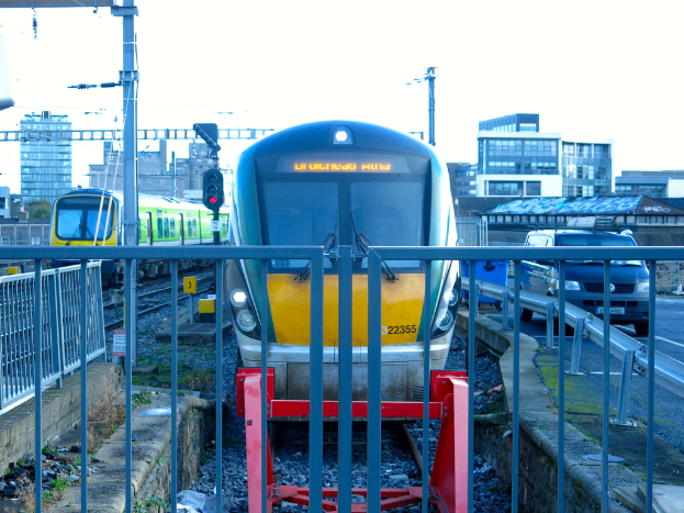 Zwei parallel verlaufende Zuggleise mit einem Zaun im Vordergrund, ein Auto auf der Stra√üe rechts daneben, eine Signalleuchte auf einem Pfahl und Geb√§ude im Hintergrund unter einem Himmel.