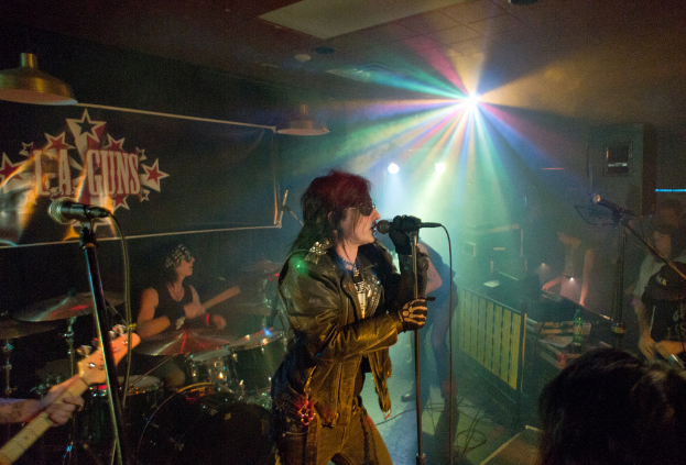 Eine Frau in einer schwarzen Jacke singt auf der Bühne in ein Mikrofon, während ein Mann mit Sonnenbrille und Stirnbinde hinter ihr Bassgitarre spielt; ein "LA Guns"-Plakat und Diskoleuchten sind im Hintergrund zu sehen.