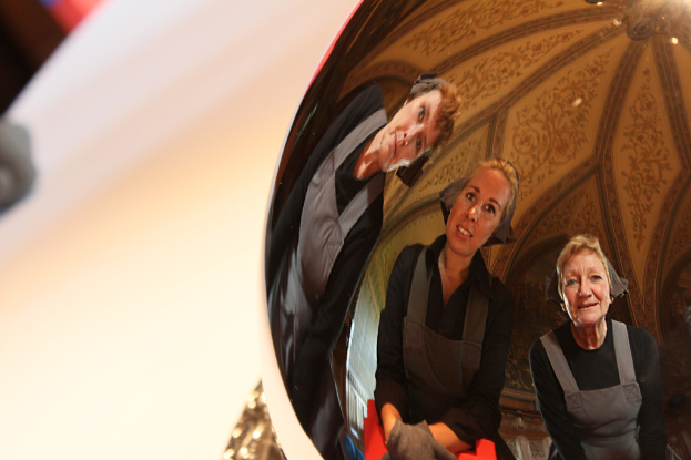 Ein reflektierender Ball auf einem weißen Tisch zeigt das Spiegelbild von drei lachenden Frauen, die leicht gebeugt sind, mit einem Dach im Hintergrund.