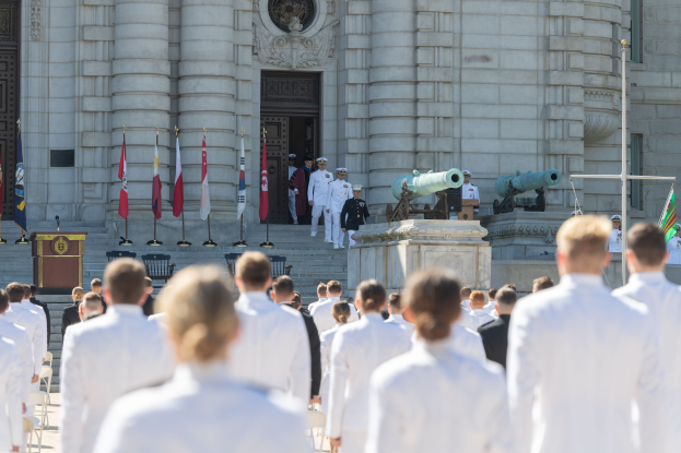 Eine Gruppe von Menschen in weißen Uniformen und Mützen steht vor einem Gebäude mit Säulen und einer Tür, mit Flaggen, einem Podium mit Mikrofon, Stufen und Kanonen im Hintergrund, was auf eine Abschlussfeier an einer Marineschule hinweist.