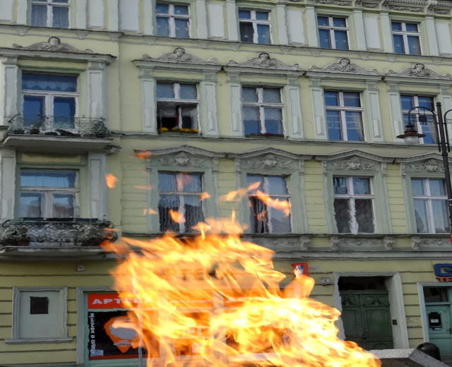 Ein großes Feuer brennt vor einem Gebäude mit Fenstern, Türen, einem Lichtmast und einer Tafel mit Text.