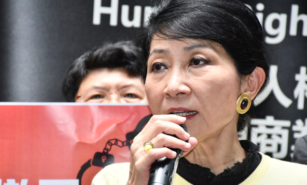 Eine Frau in einem schwarzen Kleid hält ein Mikrofon und steht selbstbewusst vor einer Menge mit einem Banner im Hintergrund, auf dem "Hong Kong Chief Executive Carrie Lam" steht.