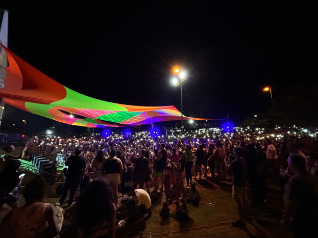 Eine große Menschenmenge steht vor einem hell erleuchteten Zelt in der Nacht während des 2018er Kunstfestivals, umgeben von farbigen Lichtern und beleuchteten Pfählen, einem Gebäude links und dem Nachthimmel im Hintergrund.