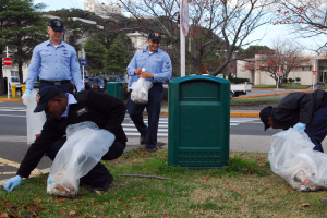 Drei Polizeibeamte in Mützen und Handschuhen sammeln M√∞ll an einer Straßenseite mit Bäumen, Gebäuden, Schildern, Fahrzeugen und einem Abfalleimer im Hintergrund an einem bewölktem Tag.