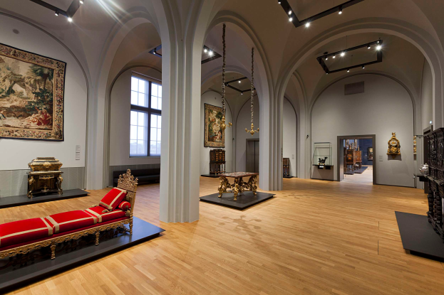 Ein geräumiger Raum im Kunsthistorischen Museum in Wien, Österreich, mit einer Couch mit Kissen, Tischen mit verschiedenen Gegenständen, Säulen, Fenstern und Deckenlampen, sowie zahlreichen paintings an den Wänden.