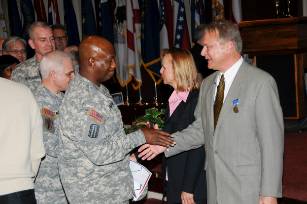 Eine Gruppe von Menschen, die mit zwei Personen Hand in Hand stehen, Flaggen im Hintergrund, ein Rednerpult mit Mikrofon und eine Topfpflanze in der Nähe.