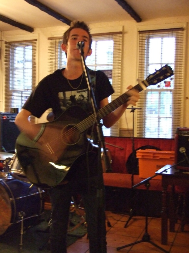 Ein Mann in schwarzem T-Shirt und Hose steht mit einer Gitarre und einem Mikrofon auf einem StГ¤nderchen, mit einer blauen Trommel links, einer Couch mit GegenstГ¤nden darauf und durchsichtigen VorhГ¤ngen im Hintergrund unter einer Decke.