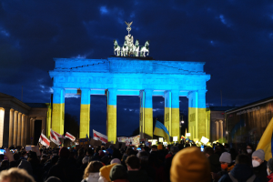 Eine Menschenmenge steht vor dem Brandenburger Tor in Berlin, hält Fahnen und Schilder in den Händen und hat rechts ein Banner mit protestbezogenem Text.