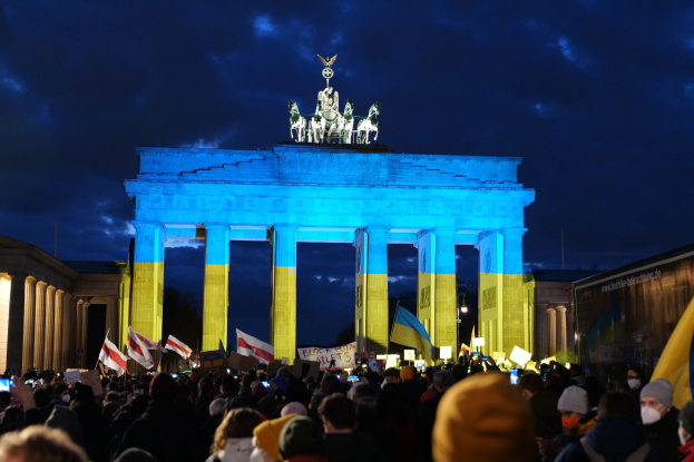 Eine Menschenmenge steht vor dem Brandenburger Tor in Berlin, hält Fahnen und Schilder in den Händen und hat rechts ein Banner mit protestbezogenem Text.