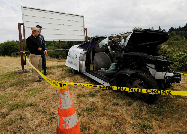 Ein Polizist in Uniform steht neben einem schwer beschädigten Auto an der Straße, mit einem Verkehrskegel und einem Band an einem Pfahl in der Nähe und Bäumen und einem bewölkten Himmel im Hintergrund.