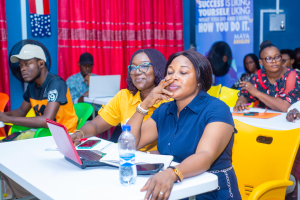 Eine Gruppe von Menschen sitzt um einen Tisch mit Laptops, Flaschen, Mobiltelefonen, Papieren, Stiften und anderen Gegenständen. Im Hintergrund ist eine Wand mit einem Vorhang und einer Fahne mit Text sowie Flaggen zu sehen.