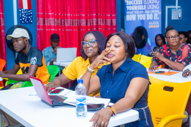 Eine Gruppe von Menschen sitzt um einen Tisch mit Laptops, Flaschen, Mobiltelefonen, Papieren, Stiften und anderen Gegenständen. Im Hintergrund ist eine Wand mit einem Vorhang und einer Fahne mit Text sowie Flaggen zu sehen.