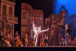 Zwei Tänzer performen auf einer Bühne im Royal Opera House in London, mit einem Publikum im Vordergrund, das auf Stühlen sitzt und steht, und einem Bildschirm im Hintergrund, der Gebäudeabbildungen anzeigt.