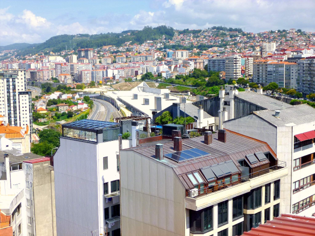 Eine Stadtansicht von einem hohen Standpunkt aus, die zahlreiche Gebäude, Bäume, Straßen, Fahrzeuge und entfernte Berge unter einem bewölktem Himmel zeigt, mit Solarpanelen auf dem Dach eines Gebäudes.